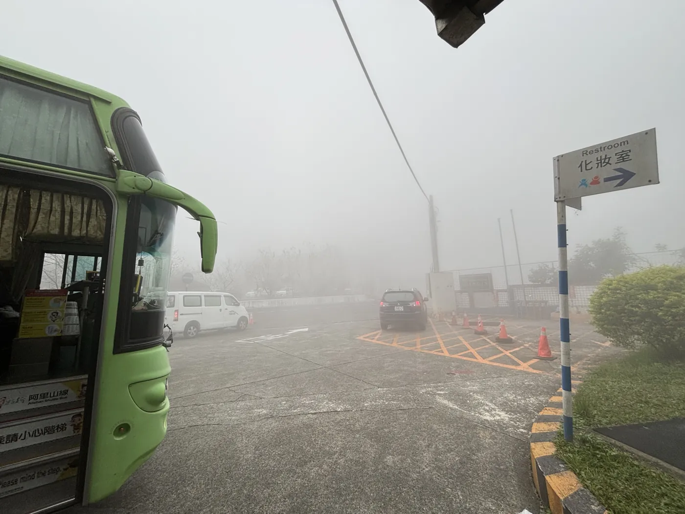 阿里山停車場，一台綠色的台灣好行阿里山線接駁公車停在濃霧中，旁邊有化妝室指標