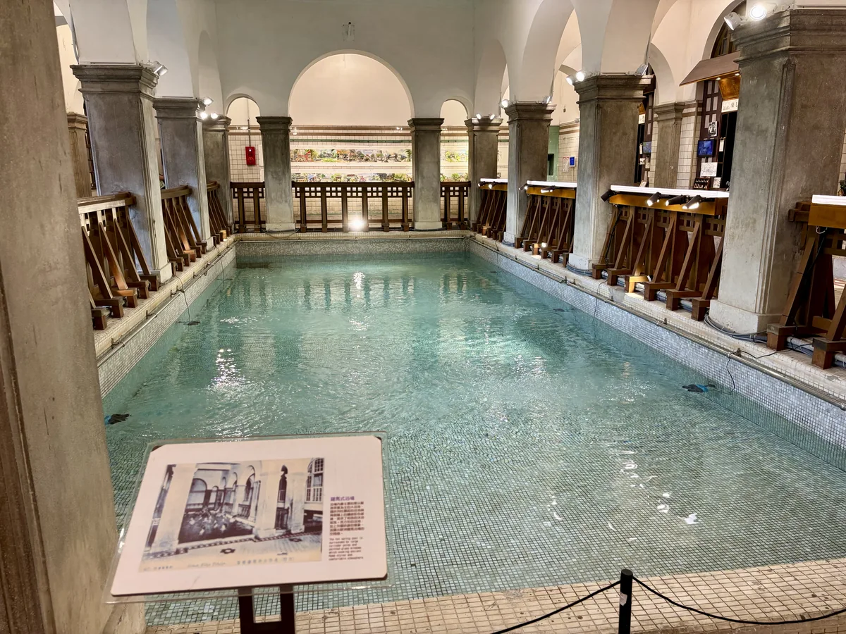 北投溫泉博物館內仿羅馬式的大浴池，池水清澈呈淺綠色，拱型迴廊環繞，池畔立著一塊介紹老照片的解說牌