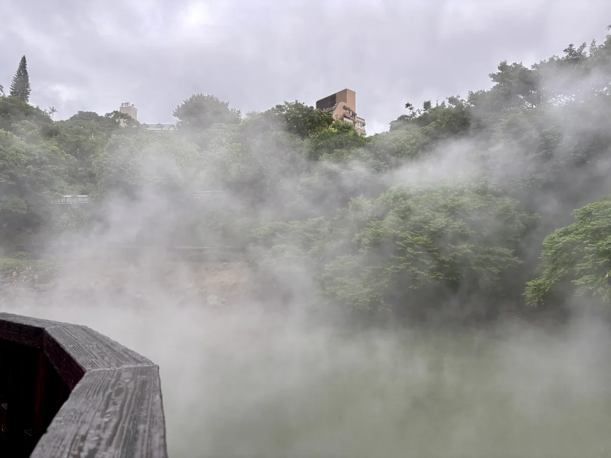 地熱谷池面白煙瀰漫，紅色木造步道欄杆延伸至池邊，遠方隱約可見建築物輪廓