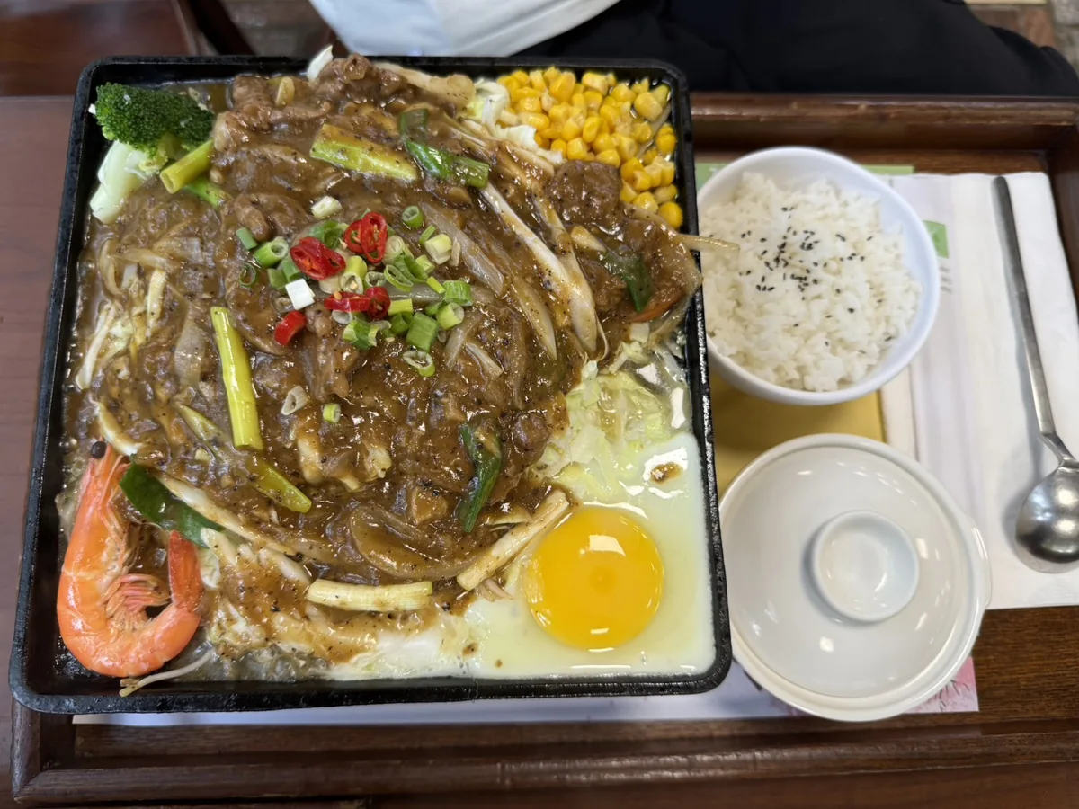 桌上擺著一鍋湯底火鍋與一份牛肉鐵板套餐，鐵板上鋪滿玉米、時蔬與一顆生蛋黃，旁邊配有白飯