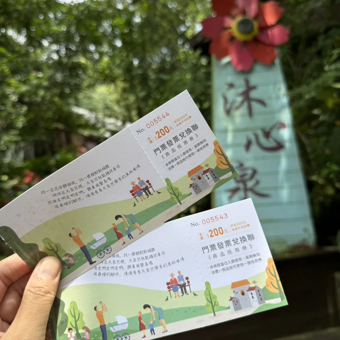 手拿兩張沐心泉休閒農場門票發票兌換聯，背景是農場入口的沐心泉招牌與紅色花朵裝飾