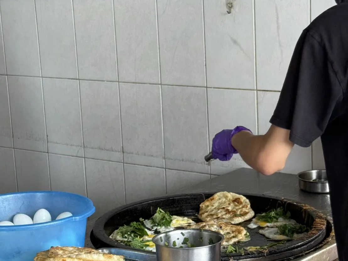 路邊攤現煎蔥油餅加蛋，師傅在大鐵盤上料理，一旁疊著剛煎好的餅