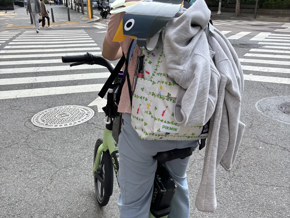 一位參加者頭戴皮克敏紙帽、背著活動束口袋，牽著淺綠色首爾公共自行車停在汝矣島的路口等紅燈