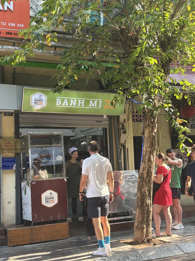 Bánh Mì 25 的店門口，綠色 BANH MI 招牌下有一台餐車，旅客在旁邊排隊